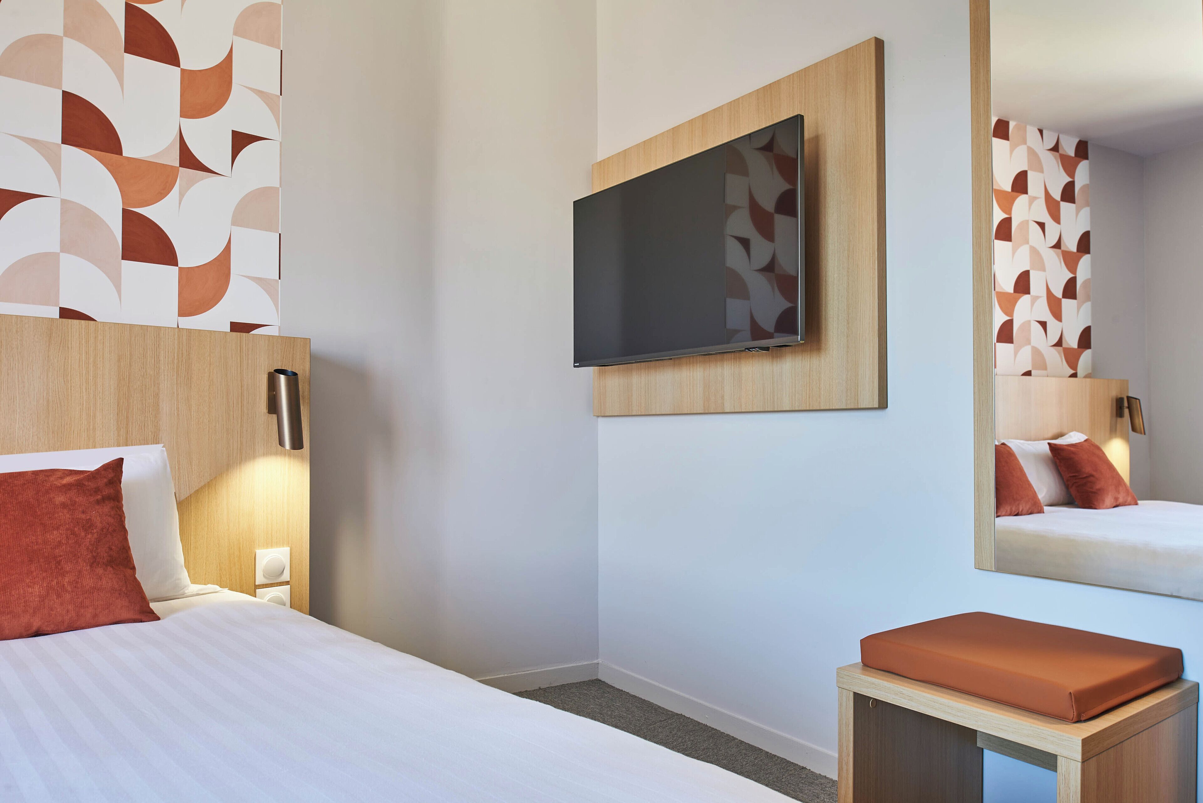 Una habitación de hotel con una cama, un televisor montado en la pared, una mesita de noche y un espejo que refleja la cama.
