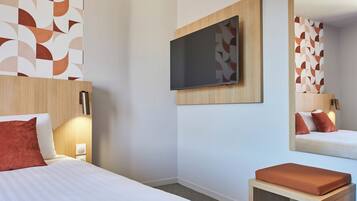 Una habitación de hotel con una cama, un televisor montado en la pared, una mesita de noche y un espejo que refleja la cama.