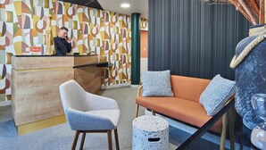 A modern hotel lobby with a wooden reception desk, a blue armchair, an orange sofa with blue cushions, and a decorative vase. - Logis Hôtel Larivaut, Puy-Guillaume (Puy-Guillaume)