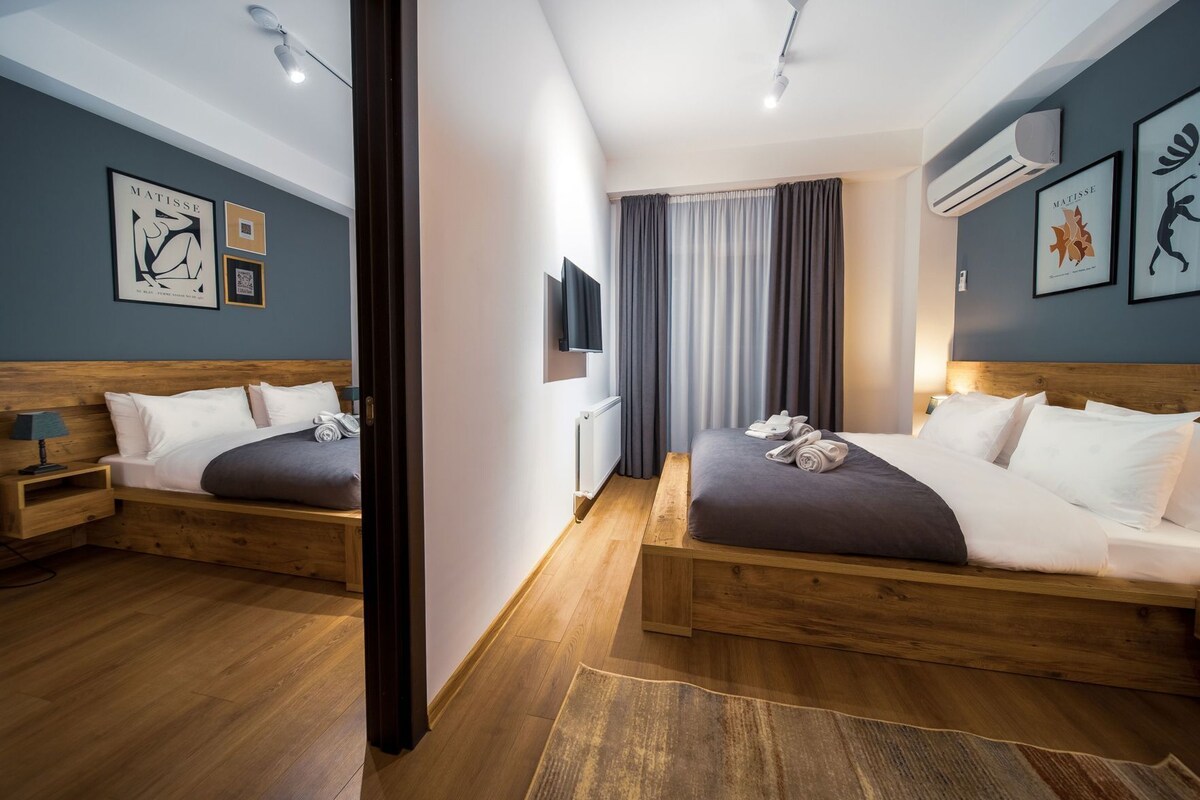 A modern hotel room with two wooden beds, a large window with curtains, and artwork on the walls.