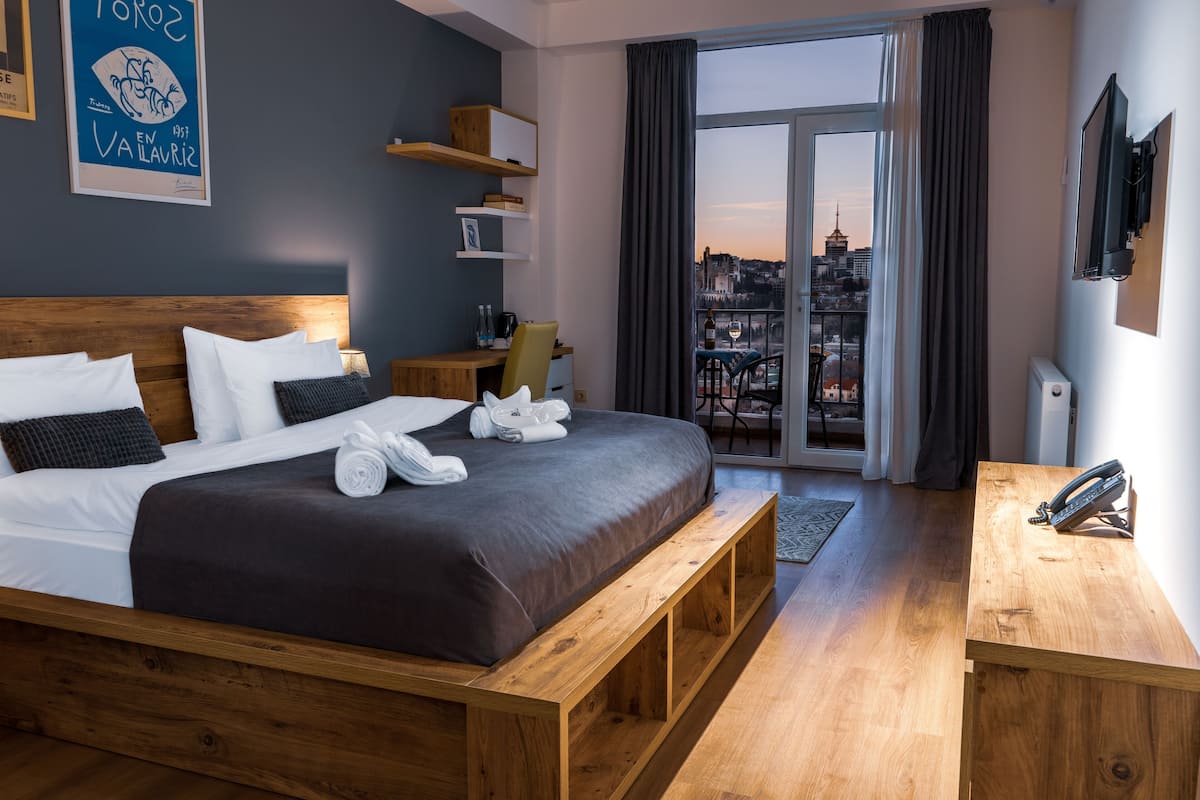 A hotel room with a large bed, a wooden bench, a desk, a TV, and a city view through the window.