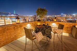 Une terrasse sur le toit, avec des chaises en osier, une table recouverte d’une nappe à motifs et des lanternes. Un arbre se dresse au centre, et on aperçoit le panorama de la ville en arrière-plan.