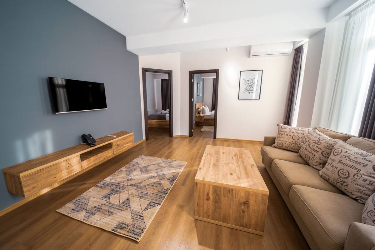A modern living room with a flat-screen TV, wooden furniture, a patterned rug, and a sofa with decorative pillows.