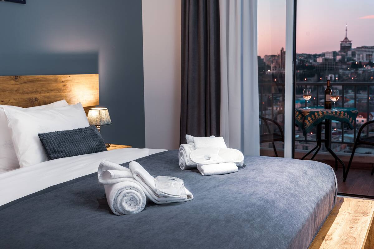 A hotel room with a large bed, a view of the city, and a balcony with a table and chairs.