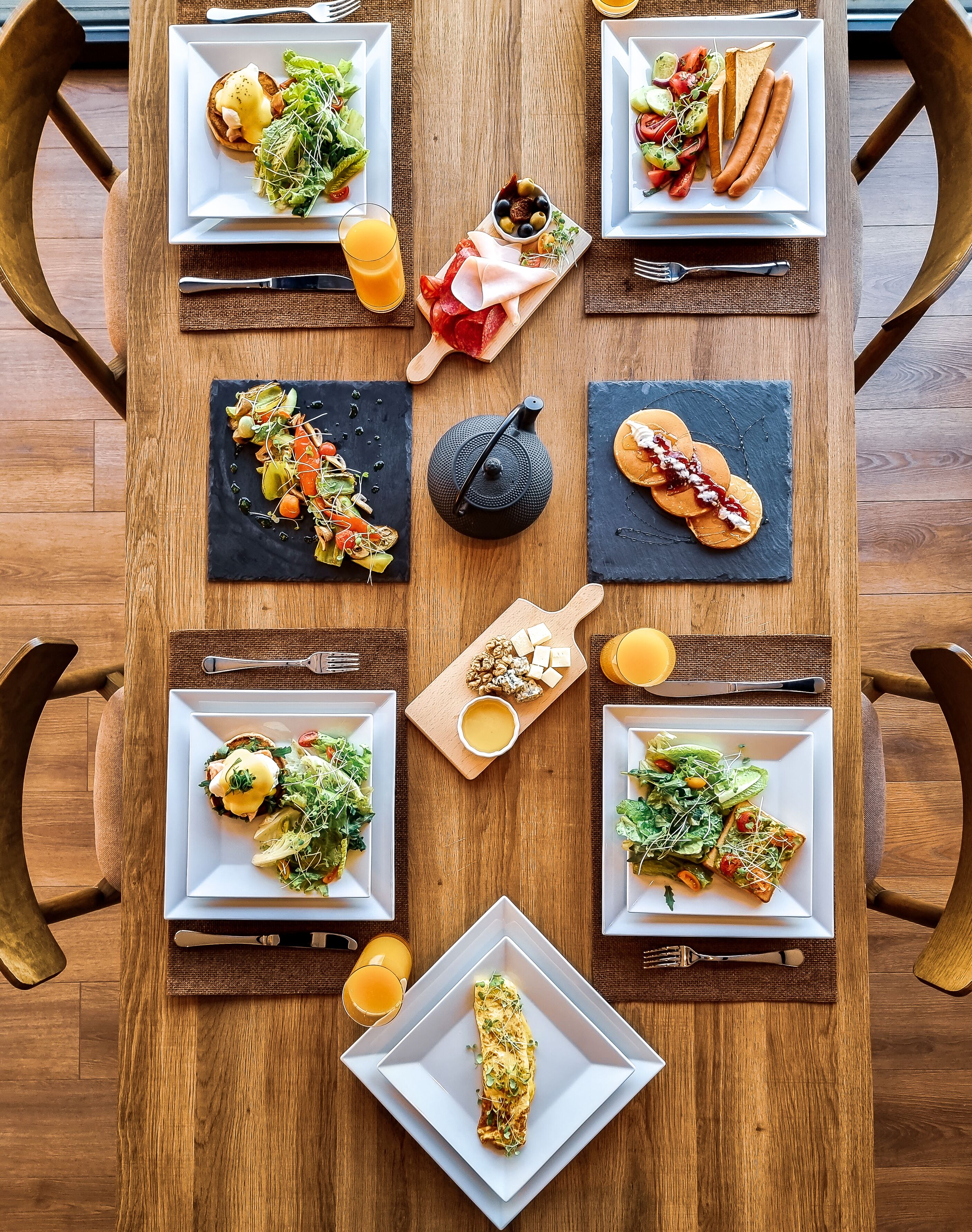 A wooden dining table with six square plates of food, a black teapot, and a wooden cutting board with cheese and olives.