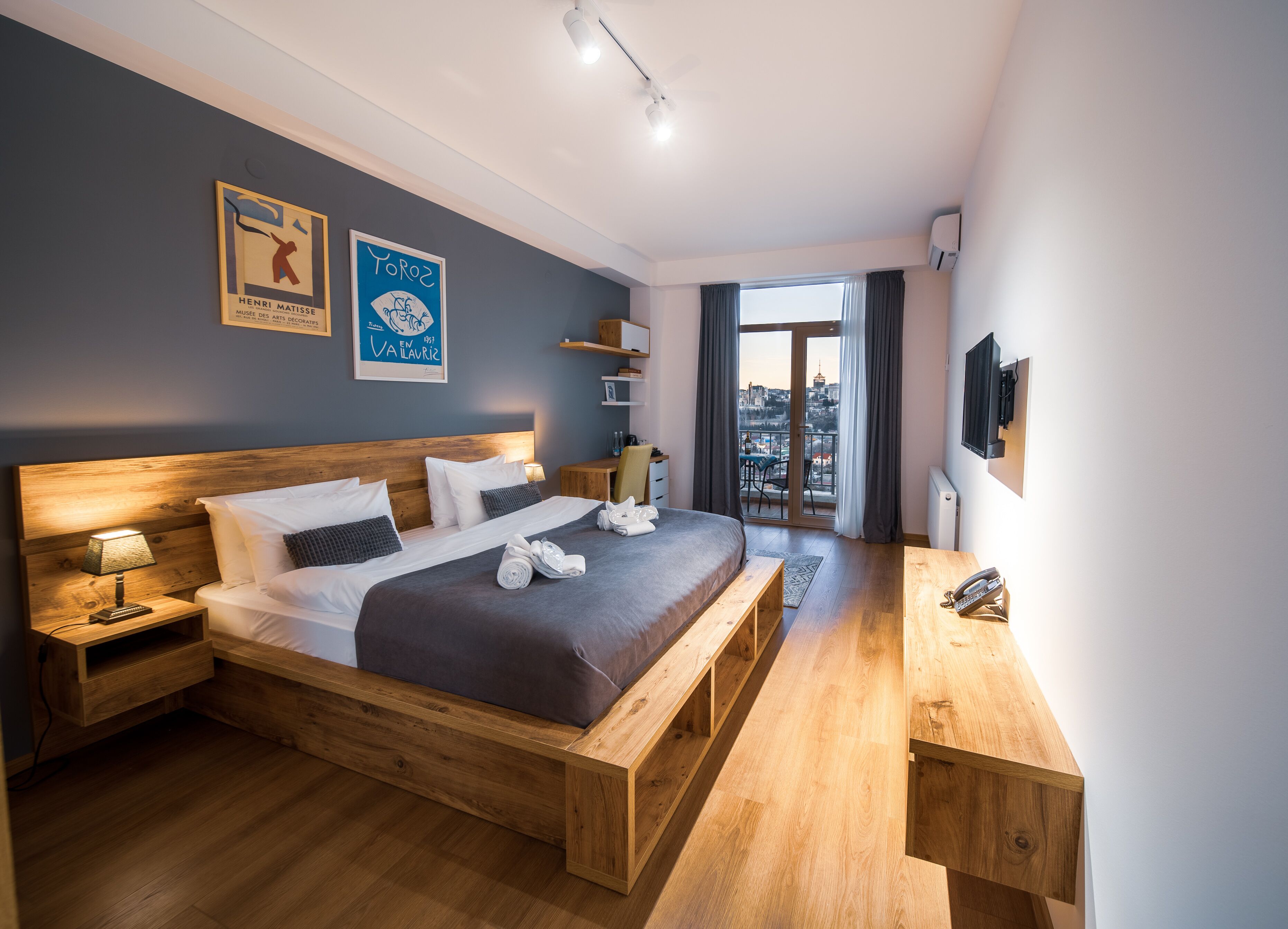 A modern hotel room with a wooden bed, a bedside table, a desk, and a balcony with a city view.
