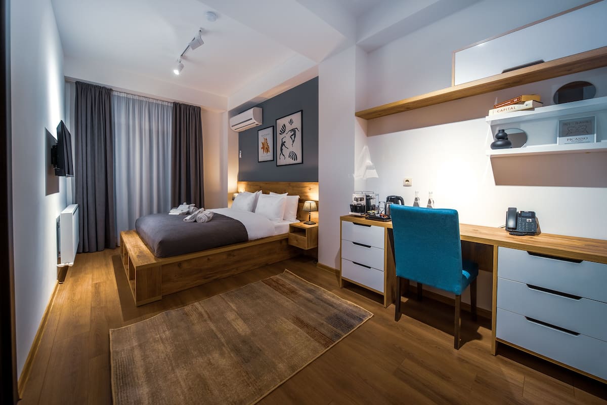 A modern hotel room with a wooden bed, a desk with a chair, a small shelf with books, and a wall-mounted TV.