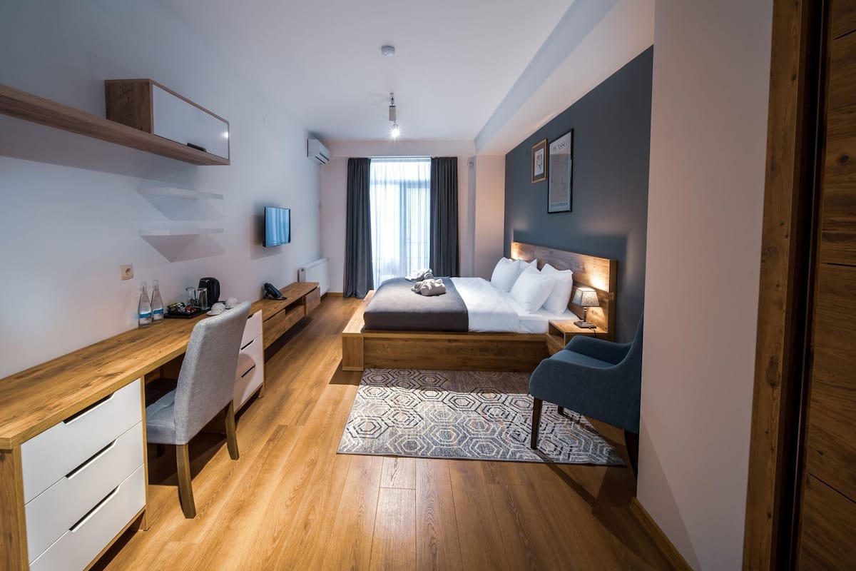 A modern hotel room with a wooden desk, a bed with white linens, a blue chair, and a patterned rug.
