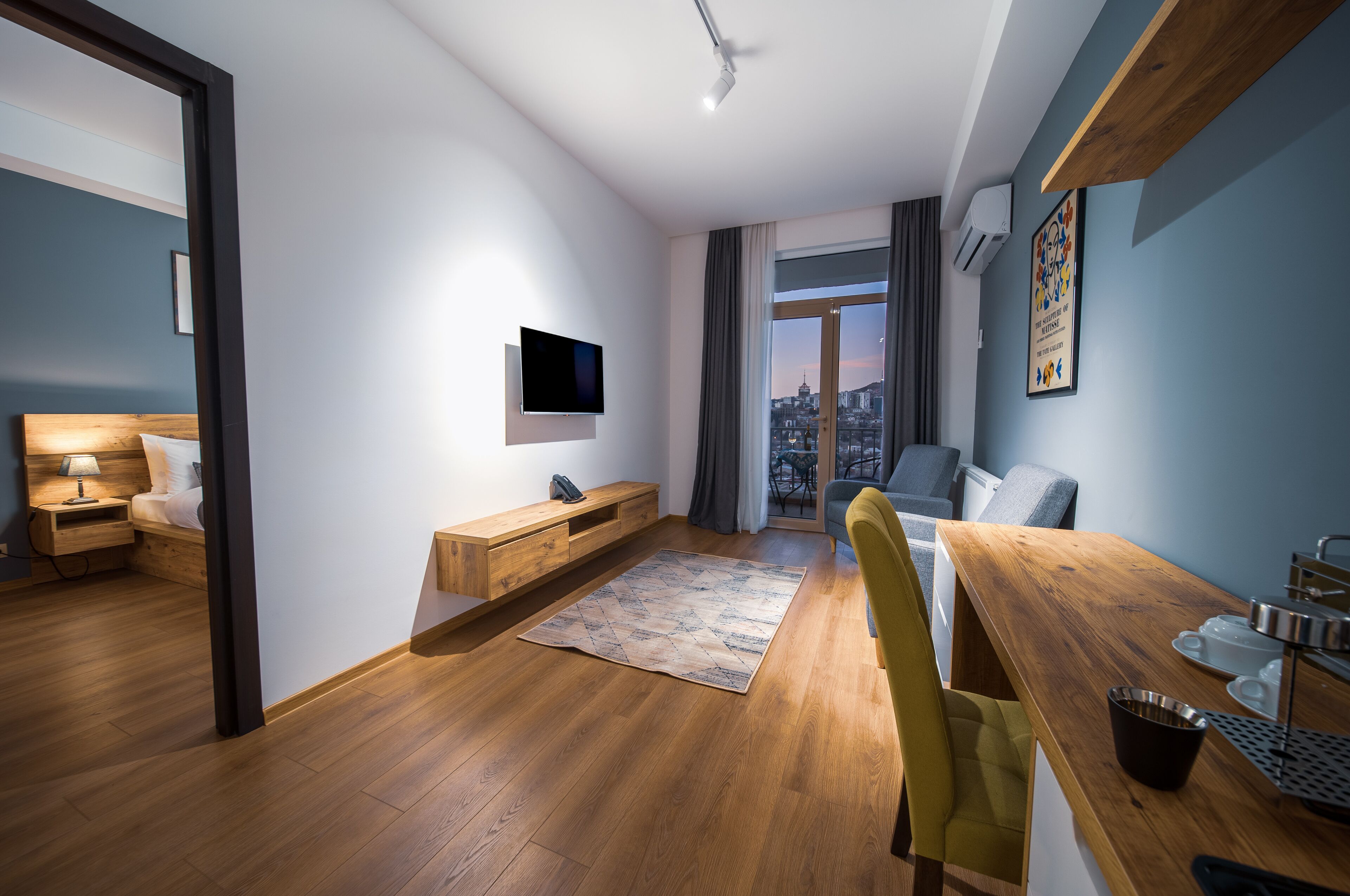 A modern hotel room with a wooden floor, a flat-screen TV, a dining table with chairs, a large window with a city view, and a small kitchenette.