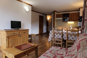 A cozy living room with a television, wooden furniture, a dining area with a table and chairs, and a kitchen in the background.
