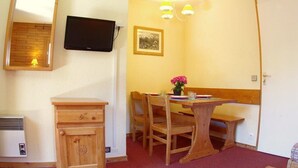 A small room with a wooden table, two chairs, a bench, a television mounted on the wall, a small cabinet, and a framed picture on the wall.