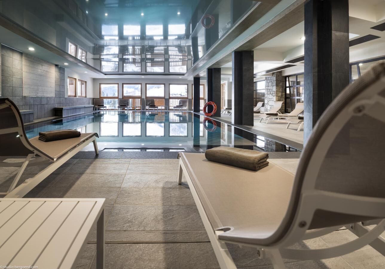 Un espace piscine intérieure moderne, doté de chaises longues et d’un mur en verre.