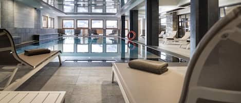 Un espace piscine intérieure moderne, doté de chaises longues et d’un mur en verre.