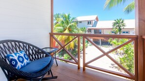 A balcony with a wicker chair and a cushion, overlooking a beach with palm trees and a building. - Casa Coral 2 Bedroom Home by RedAwning (Roatan)