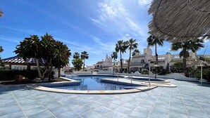 A swimming pool area with tiled flooring, palm trees, and a thatched umbrella. - Botanical 2 Bedroom Bungalow with Com Pool VM33 (Orihuela)
