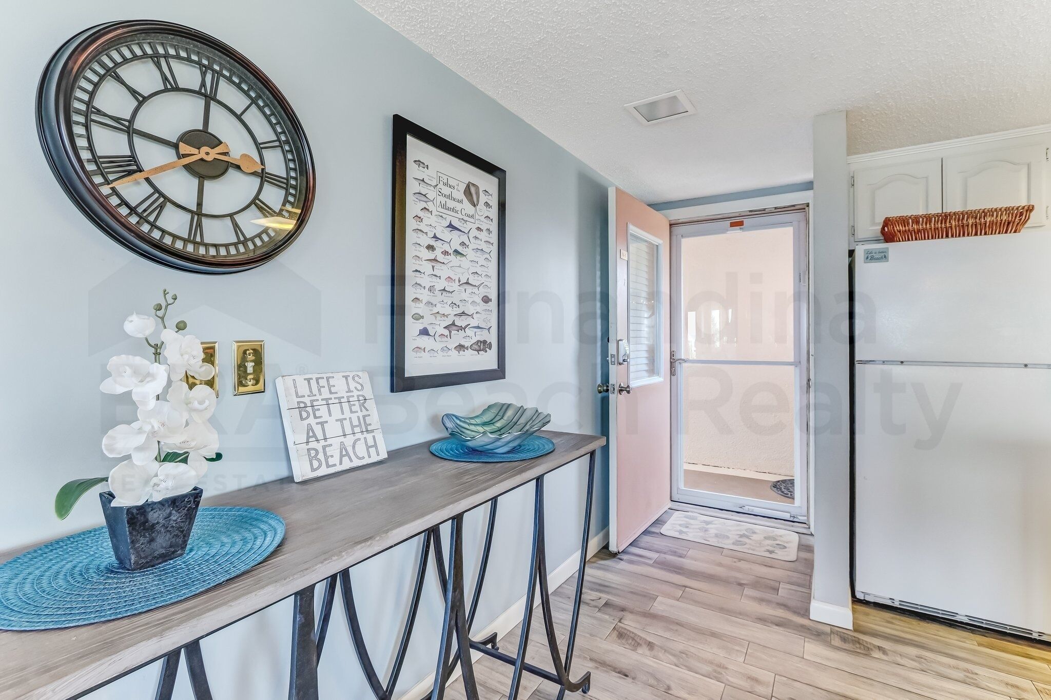 A modern kitchen with a large clock, framed fish illustrations, and a decorative bowl on a wooden table.