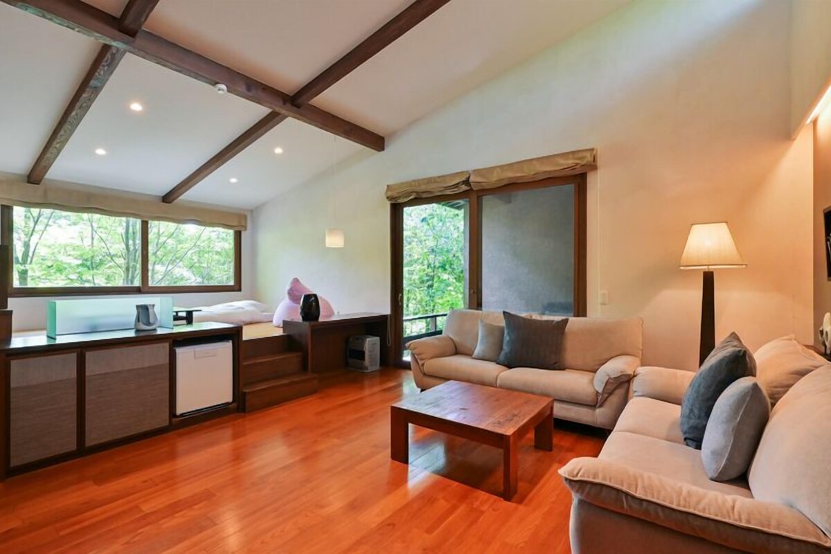 A spacious living area with a wooden floor, a sofa, a coffee table, and a desk with a chair. There are large windows providing natural light and views of greenery outside.