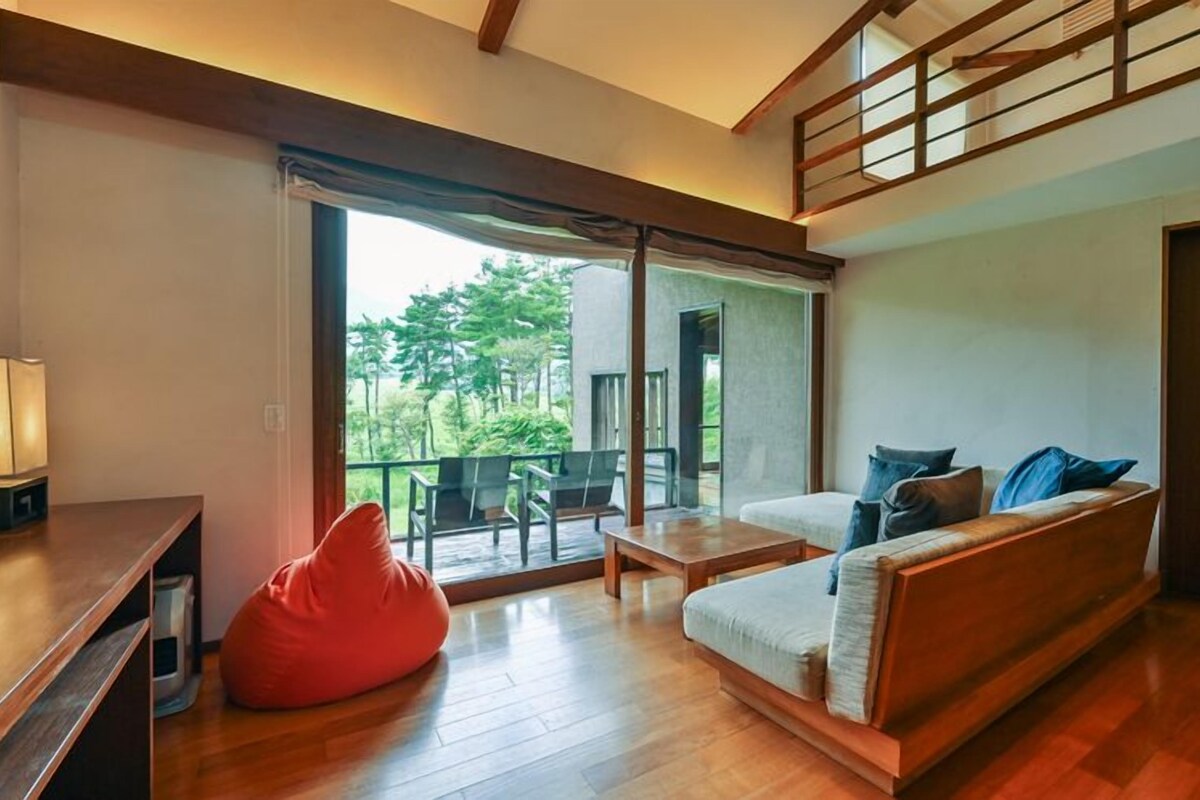 A modern living room with a wooden floor, a sofa with cushions, a red bean bag, and a balcony with a view of greenery.