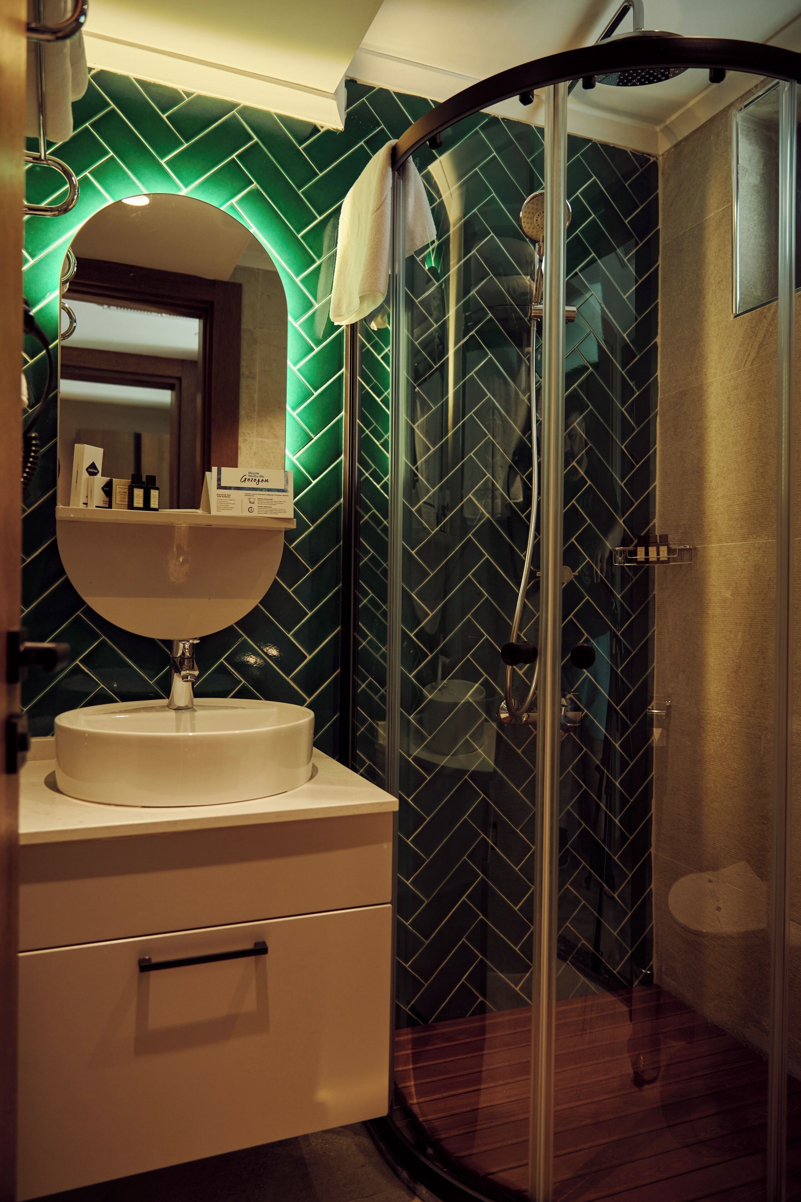Een badkamer met een glazen douchecabine, een wit wastafeltje met een spiegel erboven en een groene betegelde muur.
