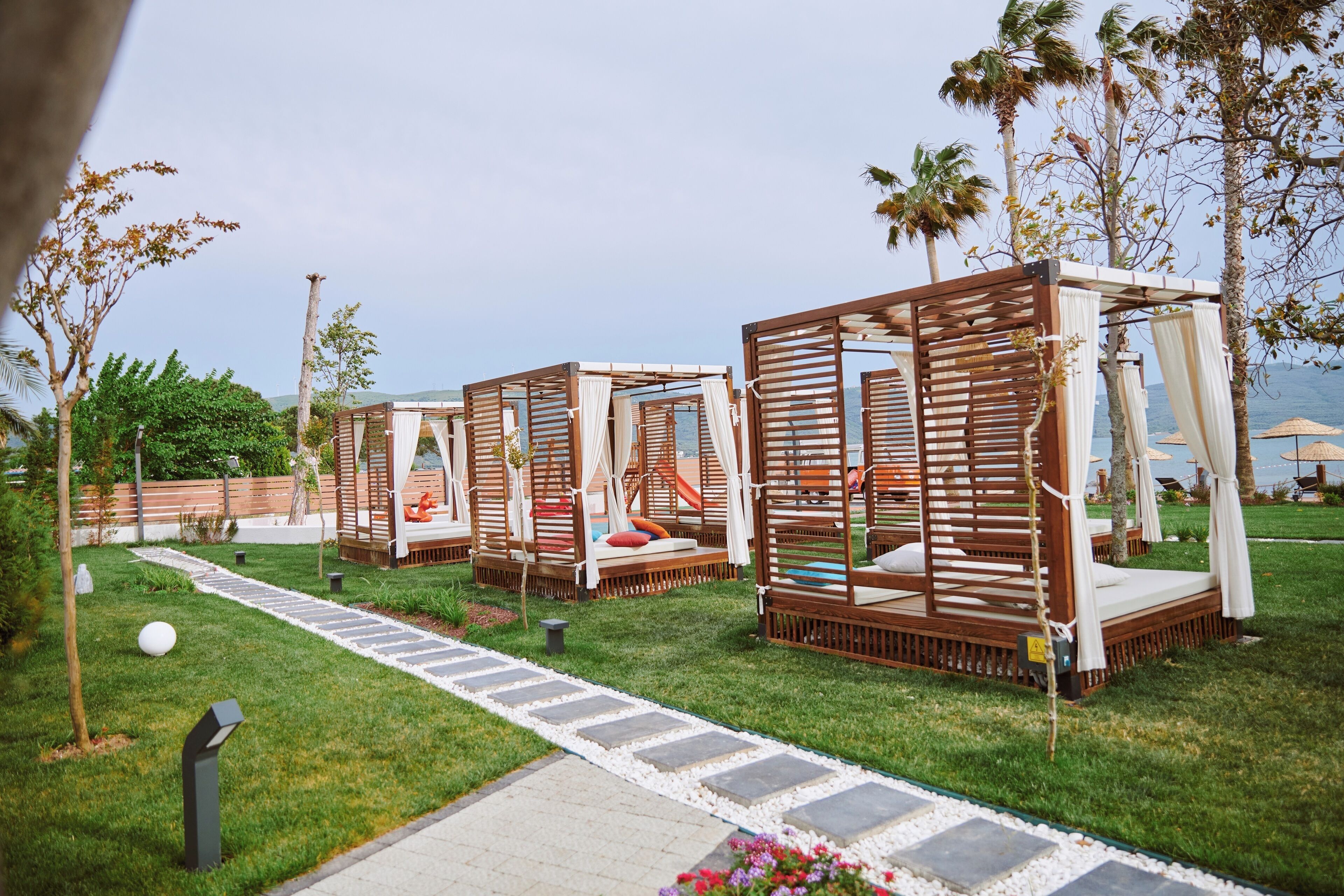 Outdoor wooden cabanas with white curtains, arranged on a grassy area with a pathway leading to them.
