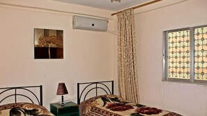 A bedroom with two beds, a nightstand, a lamp, a window with patterned curtains, and a wall-mounted air conditioner.