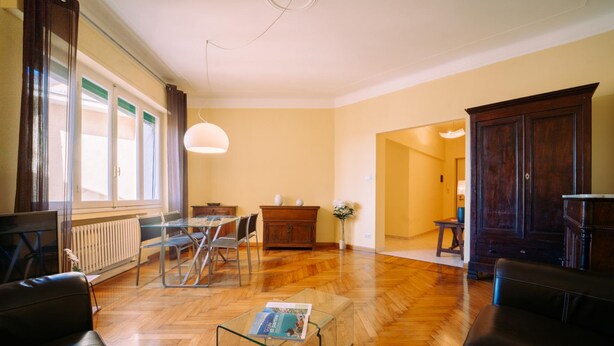 A spacious living room with a wooden floor, a glass coffee table, a sofa, a dining table with chairs, and a dark wooden cabinet. - Elegant Apartment in the Center by Wonderful Italy (Sanremo)