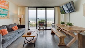 A modern living room with a grey sofa, wooden coffee table, and a wooden dining table with benches. A TV is mounted on the wall, and there is a balcony with a view of the beach. - 0311 Seaside Retreat by Atlantic Towers (Carolina Beach)