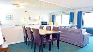 A hotel room with a dining table and chairs, a sofa, a television, and a large window with curtains. - Towers 402 2 Bedroom Condo by RedAwning (Ocean City)