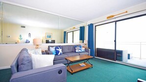 A modern living room with a grey sofa, a wooden coffee table, and a large window with blue curtains. - Towers 402 2 Bedroom Condo by RedAwning (Ocean City)