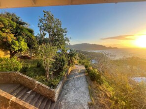 Eine steinerne Treppe führt zu einer Aussichtsplattform mit Blick auf das Meer und ferne Berge bei Sonnenuntergang.