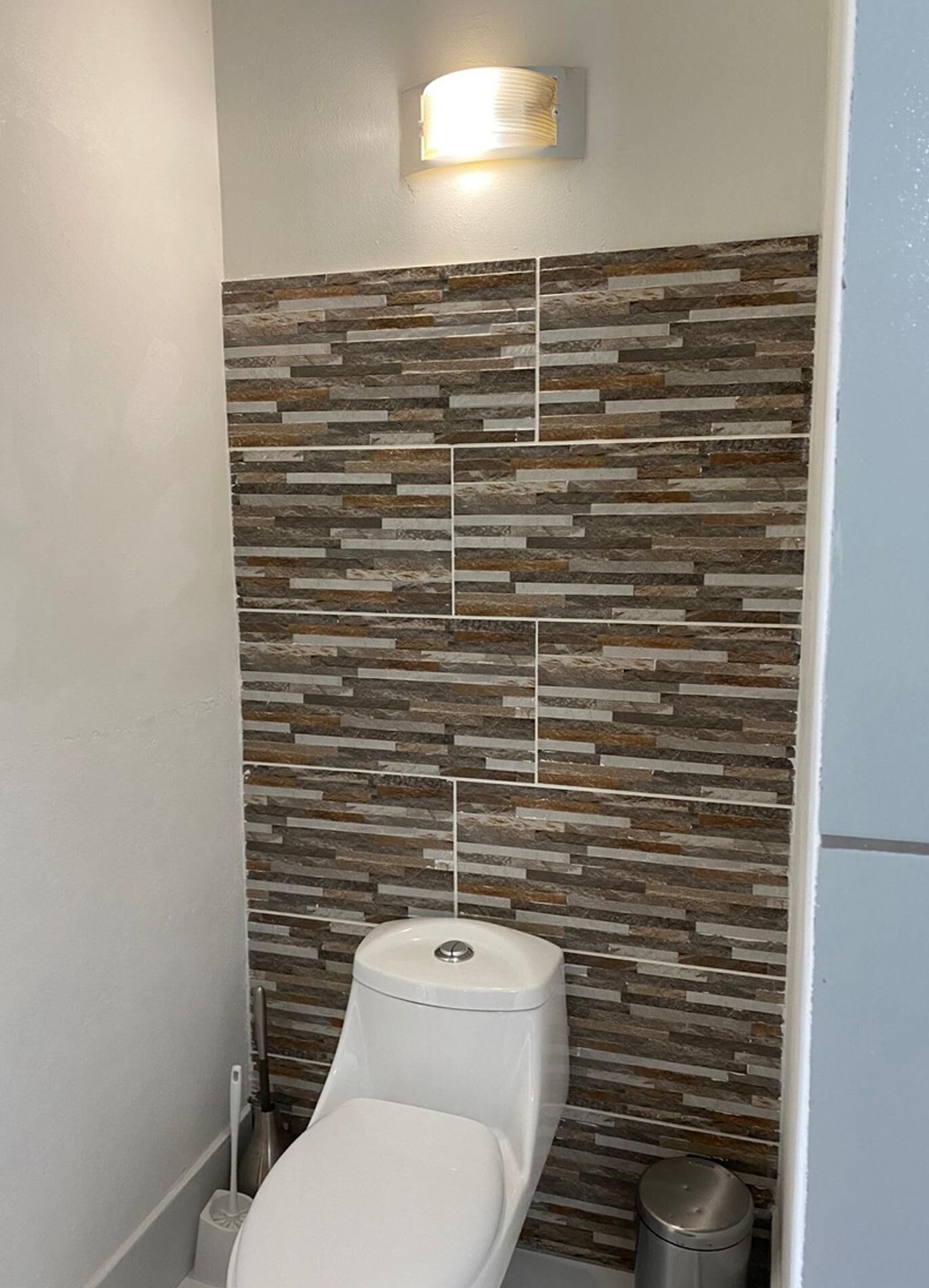 A bathroom with a stone-patterned wall, a toilet, a trash bin, and a wall-mounted light fixture.