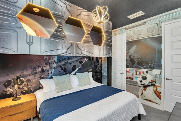 Quarto com cama, mesa de cabeceira, painel decorativo na parede e porta com arte temática espacial.