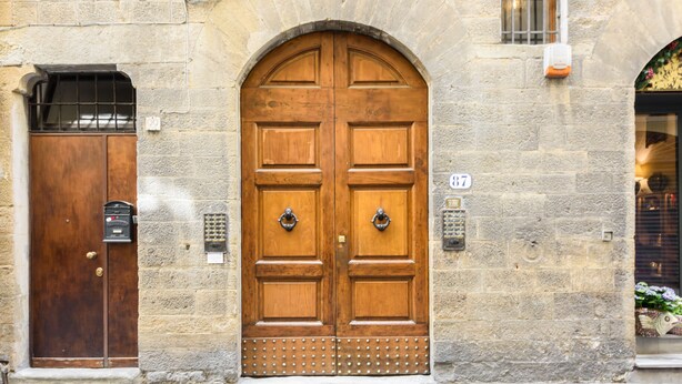 Une grande porte à deux battants en bois, ornée d’une grille métallique, une porte plus petite sur la gauche, et un mur de pierre sur lequel est apposée une enseigne au-dessus de la porte principale.