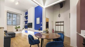Un salon moderne avec un plancher en bois, un canapé bleu, une table à manger avec des chaises bleues et un coin cuisine doté d’un comptoir blanc et d’étagères bleues.