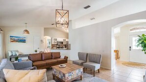 A spacious living room with a brown sofa, a grey armchair, and a coffee table with a colorful tile base. A kitchen is visible in the background, and a large window provides natural light. - Entire home, Heated Pool, Lake view and Gazebo! (Riverview)
