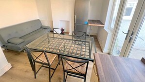 Een knus woongedeelte met een tafel met glazen blad, twee stoelen, een grijze bank, een koelkast van roestvrij staal en een houten bankje.