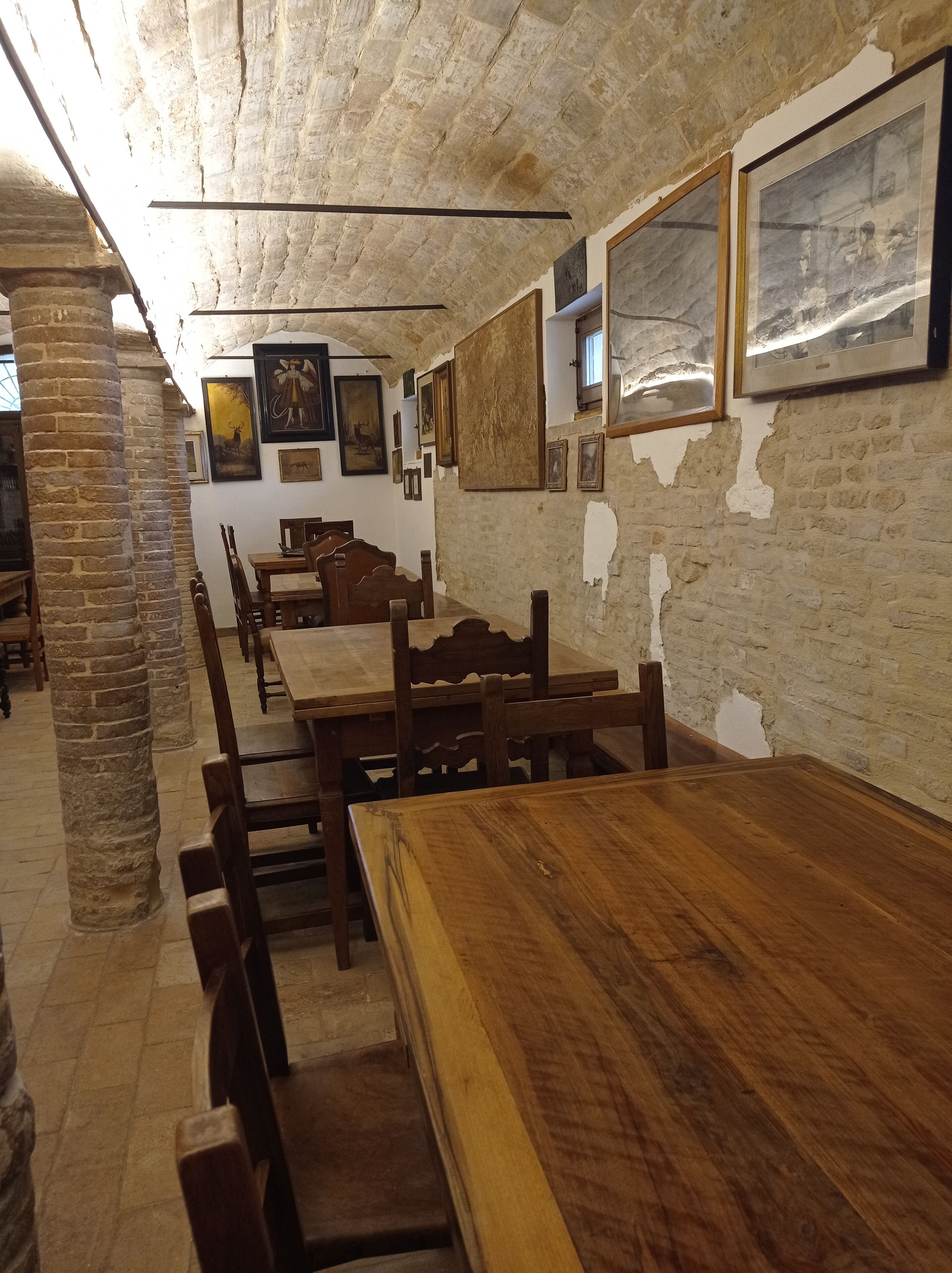 Um interior rústico com paredes de tijolo aparente, mesas e bancos de madeira, e obras de arte emolduradas nas paredes.