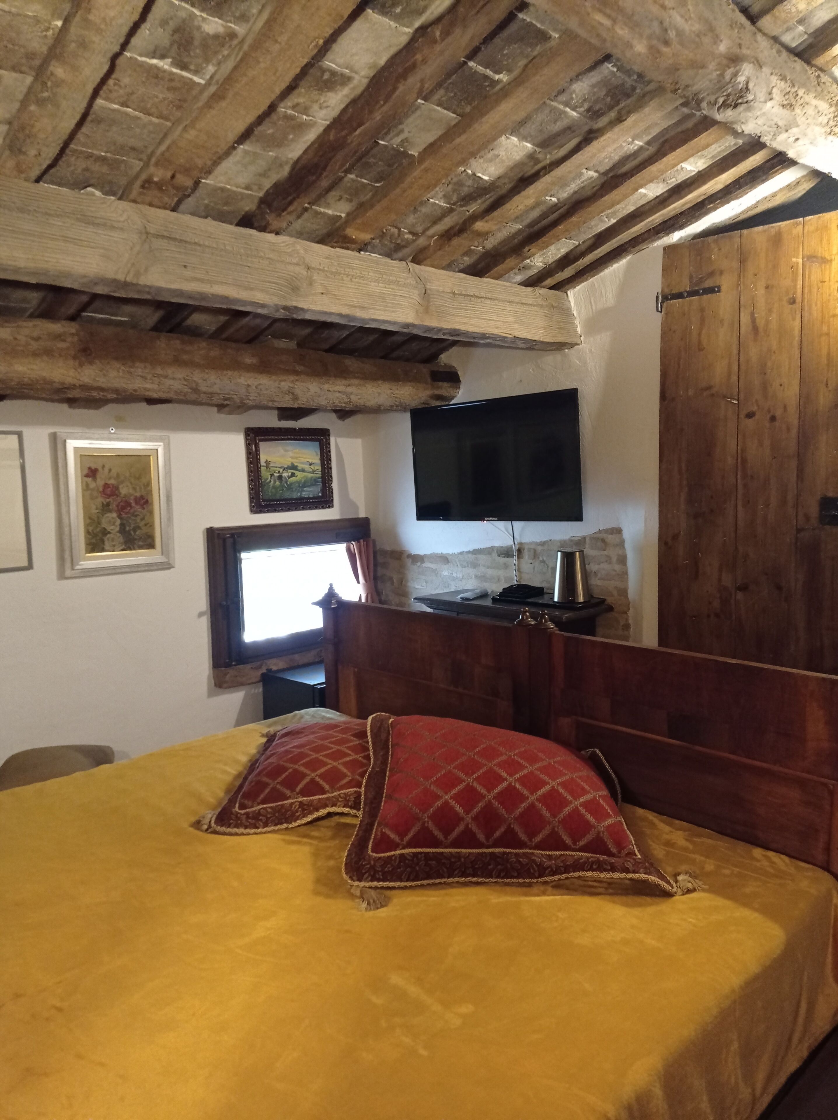 A bedroom with a wooden ceiling, a bed with yellow bedding and red pillows, a television on the wall, and a wooden wardrobe.