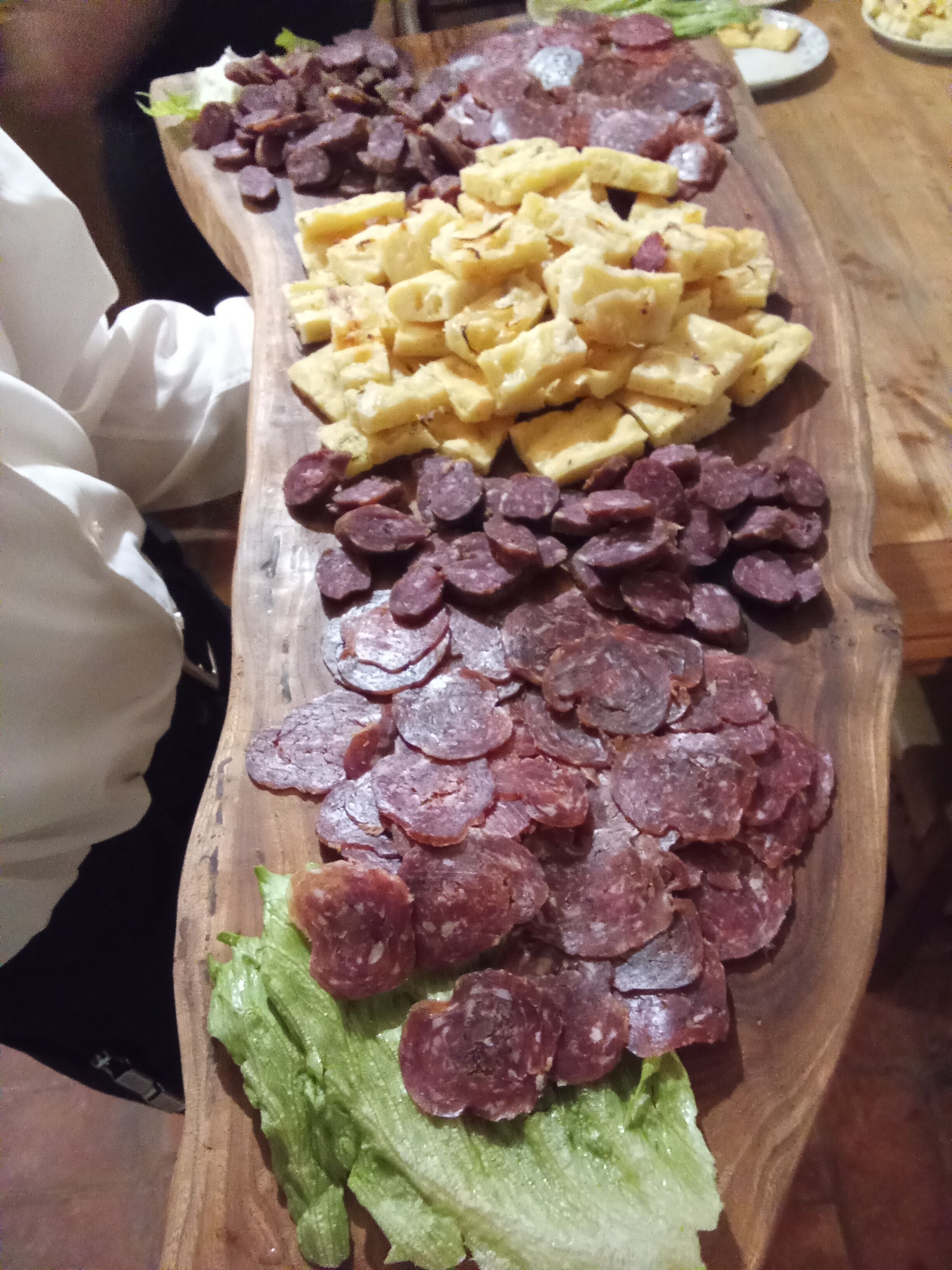 A wooden serving board with various food items including sliced sausages, a stack of waffles, and some leafy greens.