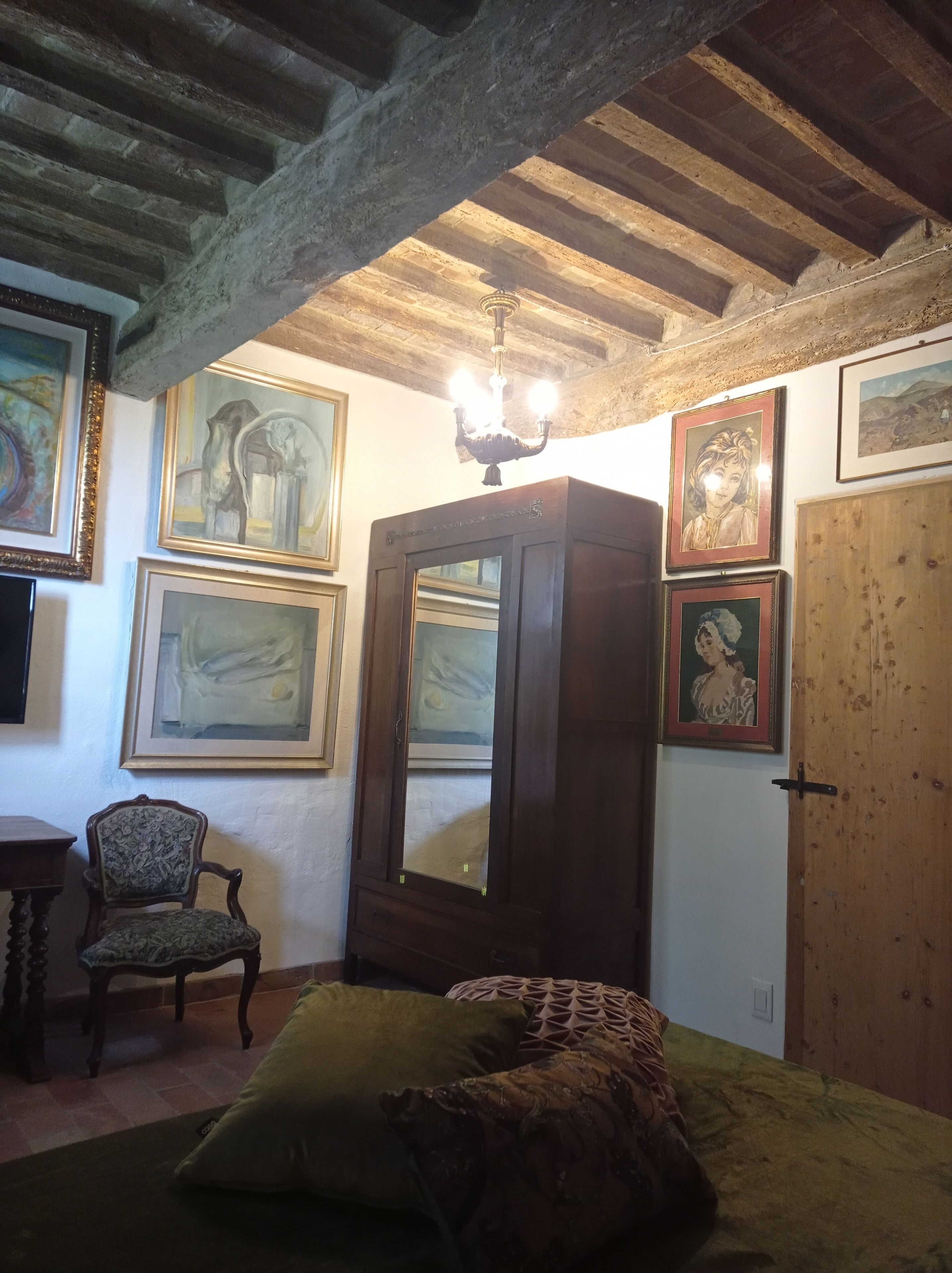 Una stanza con soffitto in legno, un letto con lenzuola verde e marrone, una sedia e diverse opere d'arte appese alle pareti.