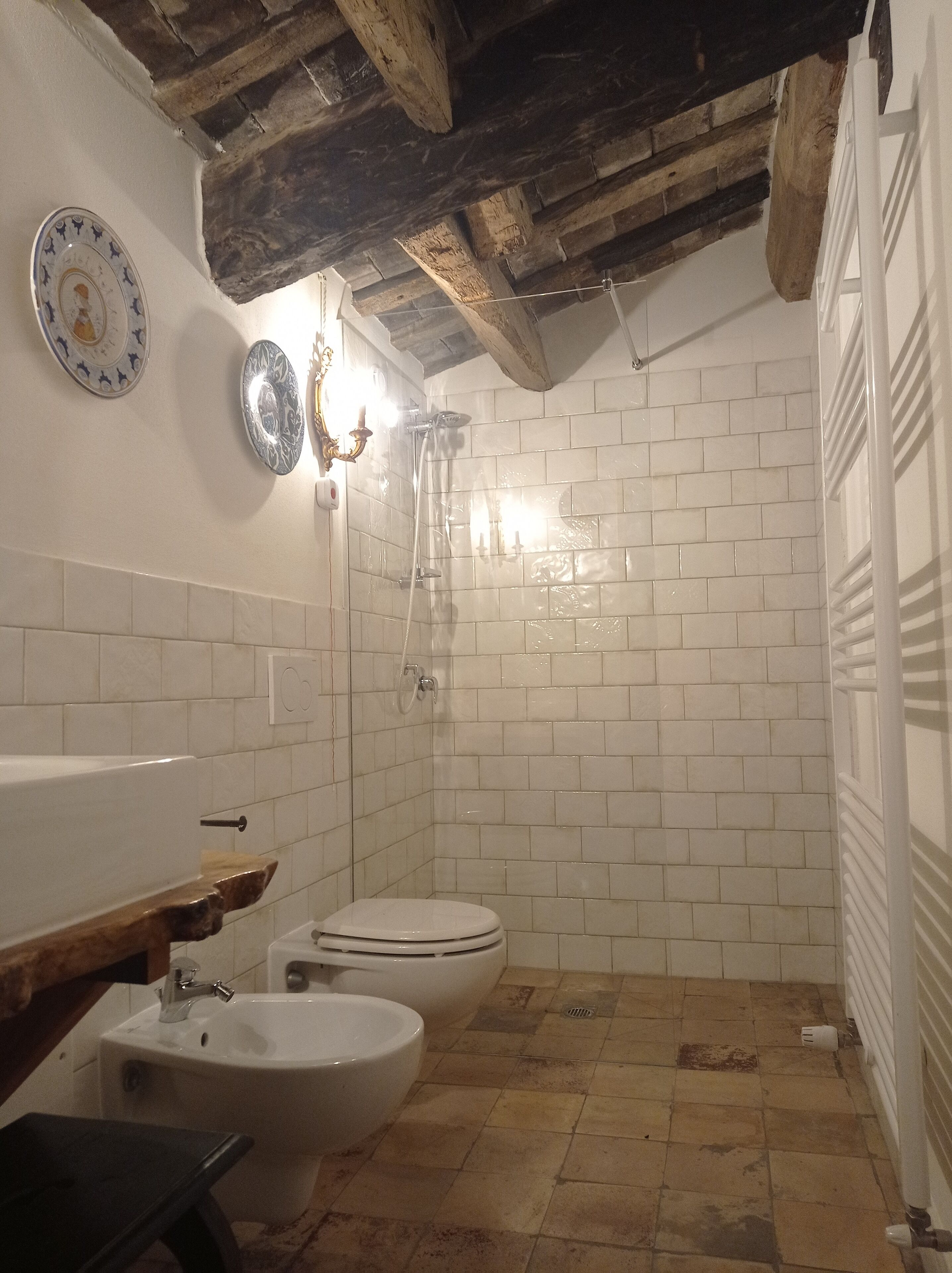 Un baño con ducha, inodoro y bidé. Presenta paredes de azulejos blancos y techo de madera.
