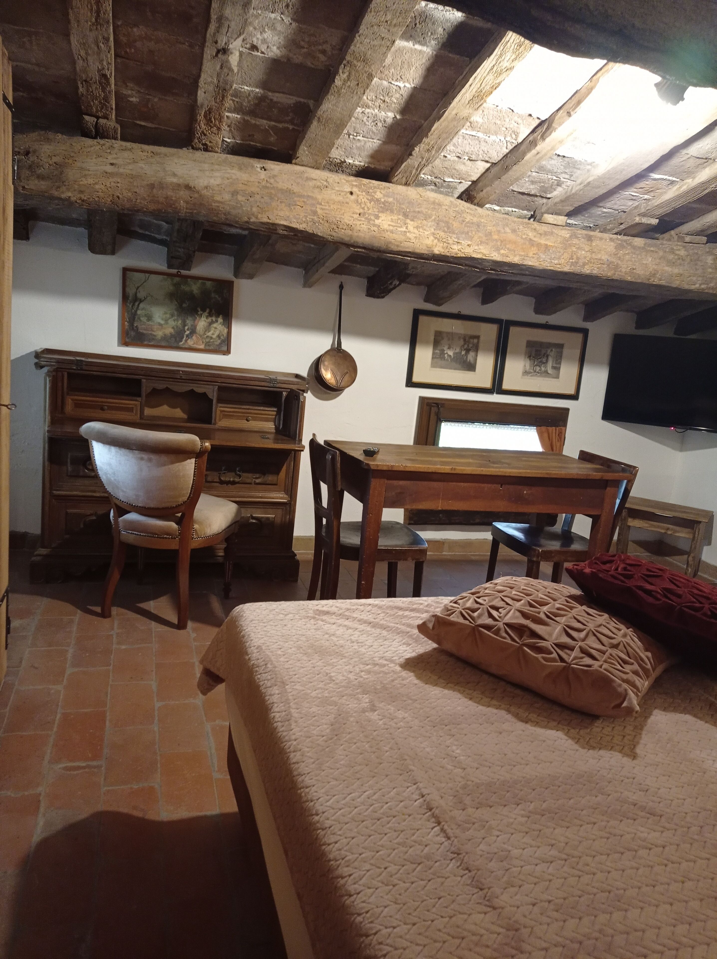 Una habitación rústica con vigas de madera a la vista, un escritorio de madera, una silla, una cama con almohada estampada y un televisor en la pared.