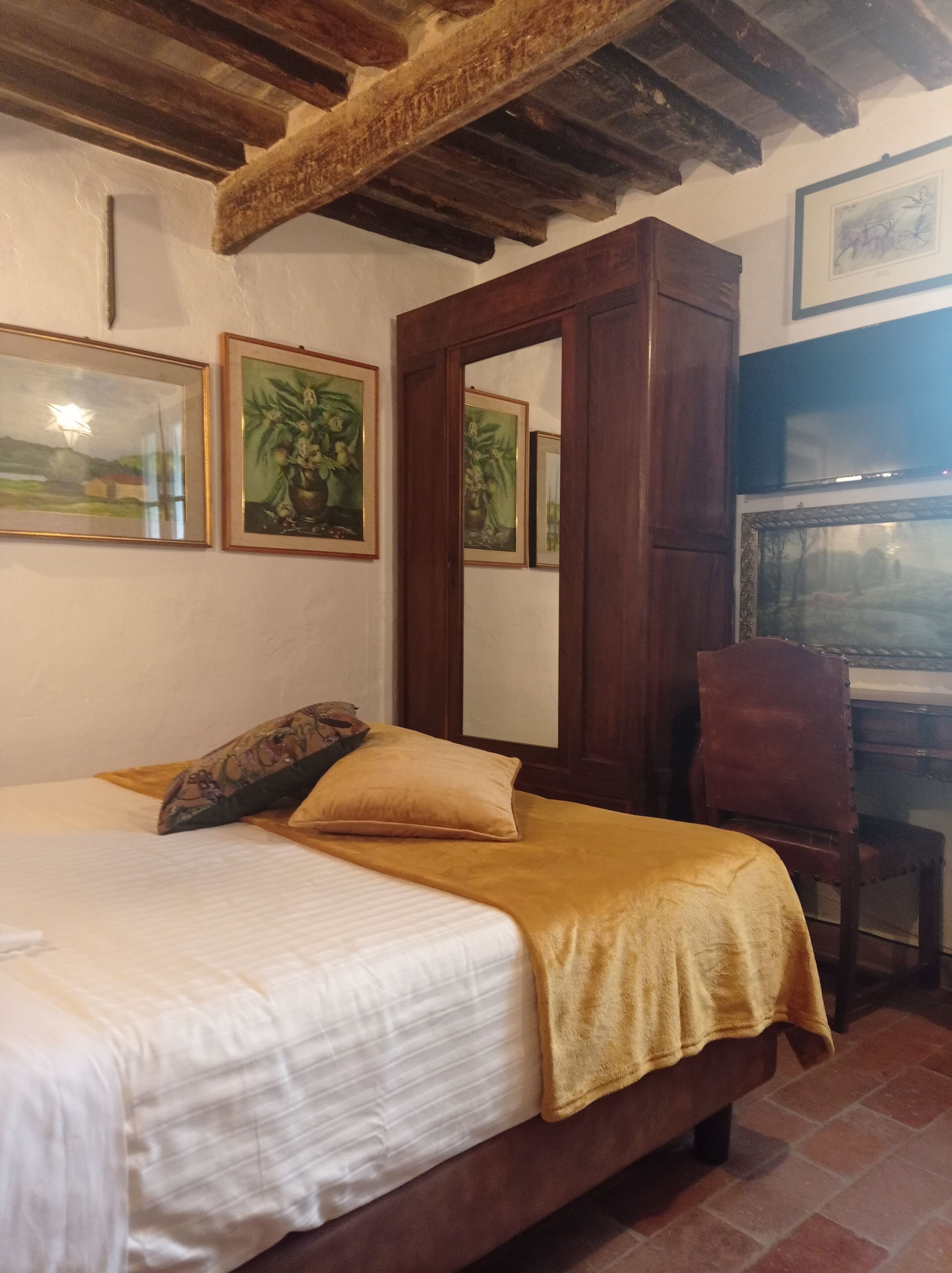 A bedroom with a bed, a wooden wardrobe, and a desk with a chair. There are framed pictures on the wall.