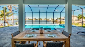 A dining area with a table set for four, overlooking a swimming pool and a lake with palm trees. - NEW - Heated Pool & Spa - Lake - Western Exposure (Cape Coral)