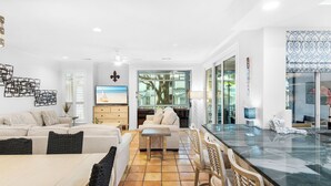 Un salon moderne avec un canapé, une table à manger et des chaises. On y trouve un téléviseur posé sur un meuble en bois, ainsi qu’une grande fenêtre donnant sur l’extérieur.