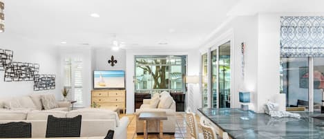 A modern living room with a sofa, dining table, and chairs. There is a TV on a wooden cabinet, and a large window with a view of the outdoors.