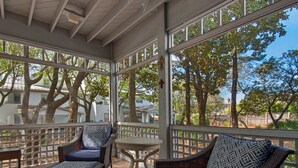 Un espace extérieur aménagé, abrité et doté de meubles en osier et d’une petite table, entouré d’une clôture blanche et de grandes fenêtres offrant une vue sur les arbres et les bâtiments.
