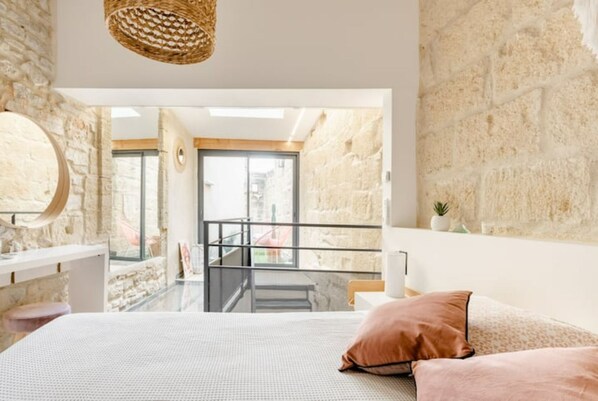 Quarto com cama, mesa de cabeceira, luminária pendente, parede de pedra e vista para o exterior.