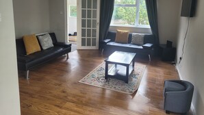 A living room with a black sofa, a wooden coffee table, a small armchair, and a TV mounted on the wall. - Beautiful 3-bed House in Ramsgate (Ramsgate)