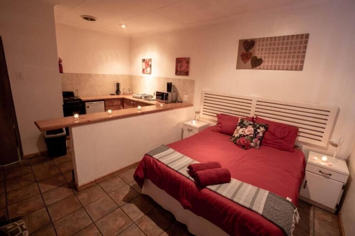 A bedroom with a bed, bedside tables, a nightstand, a small kitchen area, and a wall with heart-shaped decorations.